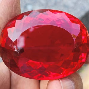 May include: A large, faceted, oval-shaped gemstone. The gem is a vibrant, deep red color, with multiple facets that reflect light. The stone is held in a hand, showcasing its size and brilliance. The overall impression is one of luxury and value.
