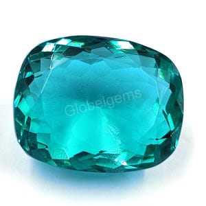 Puede incluir: Una gema grande, facetada, de color verde azulado con talla cojín. La gema tiene un alto grado de claridad y brillo, reflejando la luz a través de sus múltiples facetas. La palabra "globelgems" es visible en la superficie.