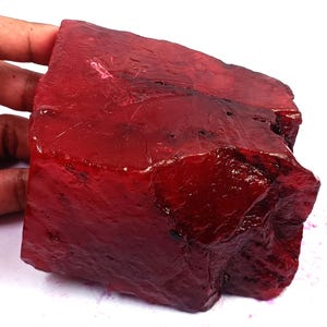 May include: A large, raw, deep red gemstone with a translucent, crystalline structure. The stone features an irregular, jagged shape with natural facets, surface pits, and a glossy, polished appearance across its uneven, blocky, and textured surface.