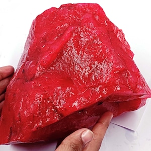 May include: A large, rough, red gemstone. The gemstone is in the shape of a rock and has a shiny, polished surface.