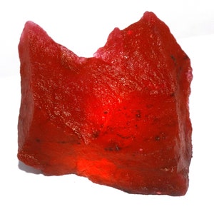 Puede incluir: Una piedra preciosa roja, cruda y translúcida, con una forma irregular y dentada y una textura cristalina rugosa. La piedra muestra diversos tonos de carmesí profundo y escarlata brillante, con reflejos de luz internos visibles.