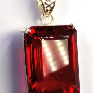 Op de afbeelding: Een rechthoekige, dieprode edelsteen hanger met een zilveren ketting. De edelsteen is facetgeslepen met een briljante slijpvorm, die het licht reflecteert. De hanger is gezet in een zilveren bezel. De ketting is een fijne, zilveren kabelketting.
