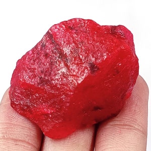 May include: A raw, unpolished ruby gemstone with a vibrant, deep red color and a textured, crystalline surface. The stone features irregular facets and natural dark inclusions throughout its structure, showcasing a translucent quality.