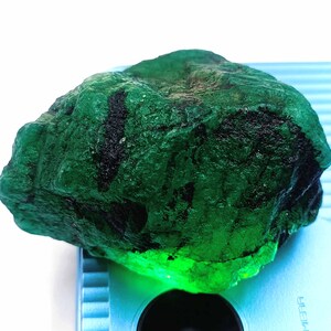 May include: A large, rough, green emerald gemstone. The emerald is illuminated from below, highlighting its deep green color.