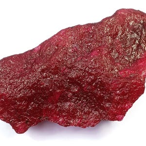 Op de afbeelding: Een ruwe, ongeslepen robijn edelsteen met een dieprode kleur. Het oppervlak van de steen heeft een textuur, met variaties in kleur en lichtreflectie. De robijn heeft een onregelmatige vorm, wat de natuurlijke, onbewerkte vorm benadrukt.