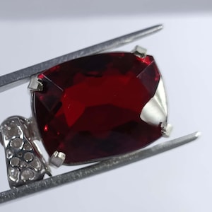 Op de afbeelding: Een vierkant geslepen, dieprode edelsteen, vastgehouden door zilveren tanden. De edelsteen is een levendig, transparant rood. Een kleiner, sierlijk zilveren element met kleine, heldere stenen is aan de edelsteen bevestigd.