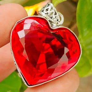 May include: A heart-shaped pendant featuring a faceted, vibrant red gemstone set within a silver-toned metal frame. The top of the pendant includes a decorative, intricate silver bail with a lattice pattern, designed for attachment to a chain.