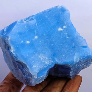May include: A large, raw chunk of bright blue mineral with a translucent, glassy texture. The stone features irregular, jagged edges and a smooth, reflective surface with subtle white inclusions scattered throughout its crystalline structure.