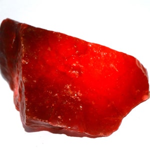 May include: A raw, irregularly shaped mineral specimen with a vibrant, translucent red colour. The stone features a jagged, crystalline texture with sharp edges and a glossy, reflective surface that catches light across its uneven, multifaceted form.
