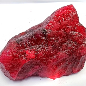 Op de afbeelding: Een ruwe, ongeslepen robijn edelsteen met een dieprode kleur. Het oppervlak van de steen heeft een textuur, met kleurvariaties en lichtreflectie. De robijn is een natuurlijk mineraal en heeft een onregelmatige vorm.