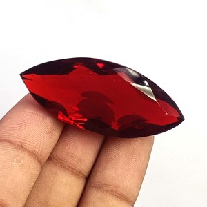 Può includere: Una gemma rosso intenso con taglio a marquise e superficie sfaccettata che riflette la luce. La pietra ha una forma ovale allungata e appuntita alle estremità, con un aspetto liscio e lucido che mostra un vibrante colore cremisi traslucido.