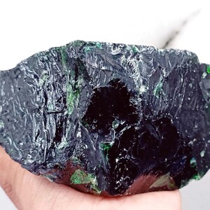 May include: A large, dark green, rough-cut gemstone with a black center. The gemstone has a textured surface and is held in a hand.