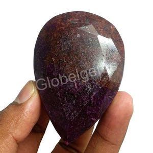 May include: A large, teardrop-shaped gemstone with a faceted surface. The stone displays a mix of deep purple and brown hues, with areas of lighter colour. The text "Globelger" is visible on the stone's surface.