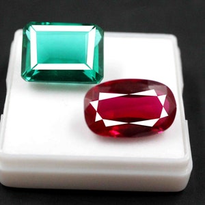 Könnte beinhalten: Zwei Edelsteine in einer weißen Schachtel. Der erste Edelstein ist ein leuchtend grüner Smaragd-Schliff-Edelstein. Der zweite Edelstein ist ein tief roter Rubin-Schliff-Edelstein.
