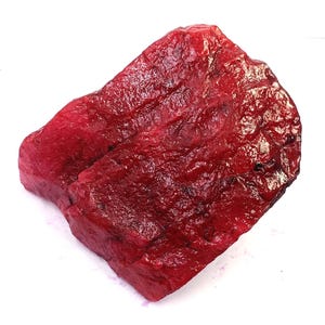 May include: A raw, unpolished ruby gemstone with a deep, saturated red colour. The stone features a rough, irregular surface texture with natural facets, ridges, and dark inclusions visible across its crystalline structure and edges.