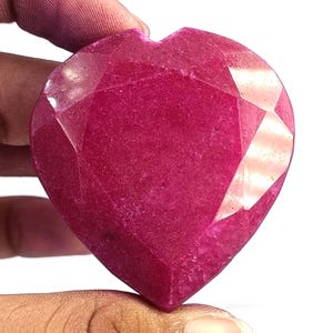 Puede incluir: Una piedra preciosa en forma de corazón con un tono magenta rosado intenso y una superficie facetada. La piedra presenta múltiples cortes geométricos que crean planos reflectantes en su parte frontal, mostrando un color vibrante y una textura pulida.