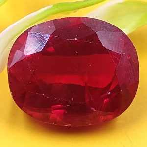 May include: A deep red, oval-shaped gemstone with a faceted surface. The stone displays a rich, saturated crimson hue and exhibits multiple geometric facets that reflect light across its polished, translucent, and slightly rounded top surface.