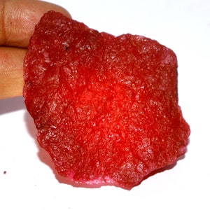 May include: A raw, unpolished gemstone with a vibrant, deep red color and a crystalline, textured surface. The stone has an irregular, somewhat flattened shape with jagged edges, displaying natural mineral formations and light-reflecting facets.