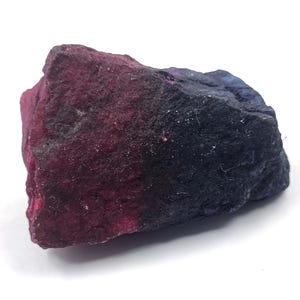 May include: A raw, unpolished mineral specimen featuring a distinct color gradient that transitions from a deep, vibrant magenta on the left side to a dark, saturated navy blue on the right side, with a rough, uneven, and textured surface.
