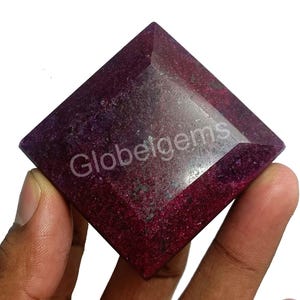 May include: A deep red square-shaped gemstone with a faceted cut. The gemstone is held in a hand and has a slight shine.
