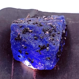 May include: A large, rough, blue gemstone with black inclusions. The gemstone is irregularly shaped and has a shiny, polished surface.