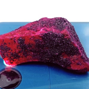 May include: A large, rough, red ruby gemstone with a dark purple hue. The ruby is on a blue surface.