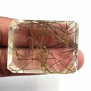 May include: A clear quartz gemstone with gold rutile inclusions. The gemstone is cut into a rectangular shape with beveled edges.