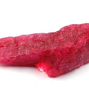 May include: A large, rough, red ruby gemstone. The ruby has a deep, rich red color and is translucent. The gemstone is unpolished and has a natural, irregular shape.