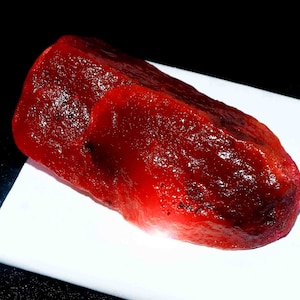 May include: A raw, unpolished ruby gemstone with a deep, vibrant red colour and a translucent, crystalline texture. The stone features an irregular, elongated shape with natural facets and a glossy surface that reflects light across its surface.