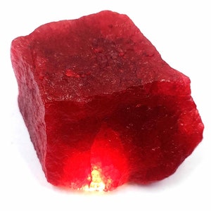 May include: A large, rough, red ruby gemstone. The ruby is opaque and has a deep red color.