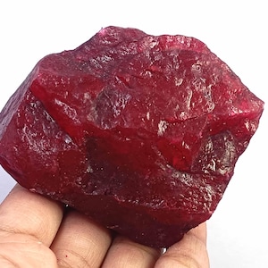 May include: A large, raw, deep red ruby gemstone with a rough, crystalline texture and irregular, angular facets. The stone displays varying shades of crimson and dark red throughout its surface, showing natural geological formations and edges.