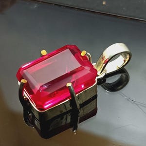 May include: A rectangular, faceted, deep pink gemstone pendant set in a metallic frame with four prongs. The pendant features a polished silver-toned bail at the top for attachment to a chain, showcasing a clear, vibrant, and translucent stone.