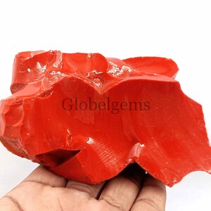 May include: A large, rough, red gemstone with a glossy finish. The gemstone has an irregular shape and is a vibrant red color.