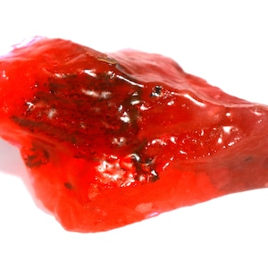 May include: A raw, translucent red mineral specimen with an irregular, jagged shape. The surface features a glossy, glass-like texture with deep crimson hues and dark, brownish-black inclusions embedded within the crystalline structure.
