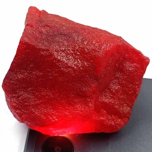 May include: A large, rough, red gemstone. The gemstone is illuminated from below, making it appear to glow.