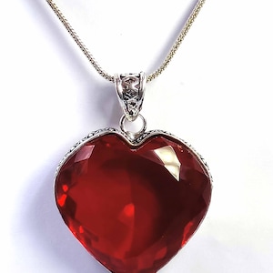 May include: A heart-shaped red gemstone pendant set in a silver frame with intricate filigree detailing along the edges. The pendant hangs from a thin silver chain, featuring a faceted surface that reflects light across its deep crimson colour.