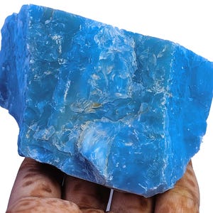 May include: A large, raw chunk of bright blue mineral with a translucent, crystalline texture. The stone features irregular, jagged edges and a glossy, reflective surface with varying shades of vibrant cyan and deep sky blue throughout the piece.
