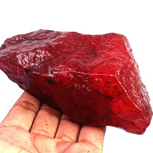 May include: A large, raw, deep red gemstone with a rough, crystalline texture and irregular, angular facets. The stone displays varying shades of crimson and scarlet, with a translucent quality that catches light across its uneven, jagged surface.