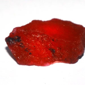 May include: A raw, unpolished gemstone with a vibrant, translucent red color. The stone features an irregular, jagged shape with a textured, crystalline surface and small, dark inclusions scattered across its bright, glowing red body.