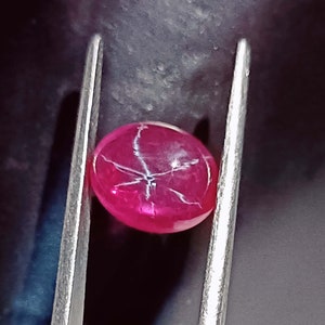 May include: A close-up of a pink star ruby gemstone with a star-shaped pattern in the center. The ruby is held in a pair of tweezers.