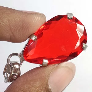 May include: A teardrop-shaped, faceted, bright red gemstone pendant with a silver-colored bail. The gemstone is held in place by silver prongs. The pendant is held by a person's fingers.