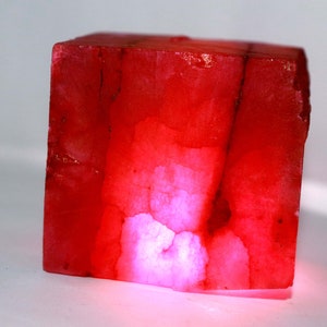 May include: A translucent, cube-shaped mineral specimen displaying a vibrant red color with internal veining and fractures. The object exhibits varying shades of crimson and pink, with a bright, glowing light source emanating from the bottom section.