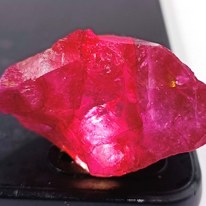 May include: A raw, uncut ruby gemstone with a vibrant, deep red hue. The natural crystal formation displays a rough, textured surface with multiple facets. The gemstone is displayed on a dark surface, highlighting its color and clarity.