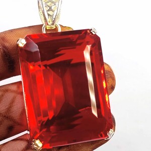 May include: A large rectangular pendant featuring a vibrant red gemstone with a faceted cut. The stone is secured by four metallic prongs at the corners and is attached to a decorative, textured silver-toned metal bail at the top.