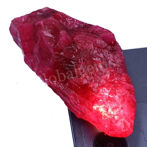 May include: A large, rough, red ruby gemstone. The ruby is translucent and has a deep red colour. The ruby is sitting on a black surface.