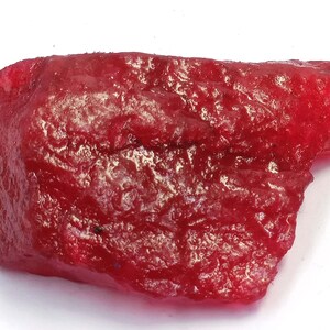 May include: A raw, uncut ruby gemstone, displaying a rich, deep red hue. The surface is textured, showing ridges and tonal variations. The ruby is presented against a white backdrop, emphasizing its natural, unpolished state.