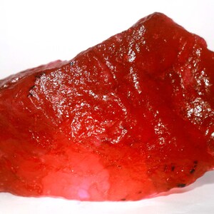 Puede incluir: Una piedra preciosa roja, cruda y translúcida, con una forma irregular y dentada y una textura cristalina. La piedra muestra tonos carmesí profundos con niveles variables de transmisión de luz en toda su superficie y bordes afilados.