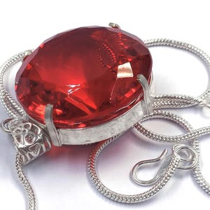 Op de afbeelding: Een zilveren ketting met een grote, ovale, helderrode edelsteen hanger. De hanger is gezet in een zilveren bezel en hangt aan een fijne slangenketting. De sluiting is een eenvoudige haak-en-oog sluiting. De edelsteen is gefacetteerd.