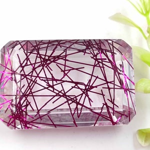 May include: A clear, faceted gemstone with a pink, web-like pattern throughout. The gemstone is cut in a rectangular shape.