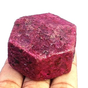 May include: A raw, hexagonal ruby crystal with a deep magenta and crimson colour palette. The surface of the gemstone displays a rough, unpolished texture with visible crystalline facets and dark mineral inclusions throughout the structure.